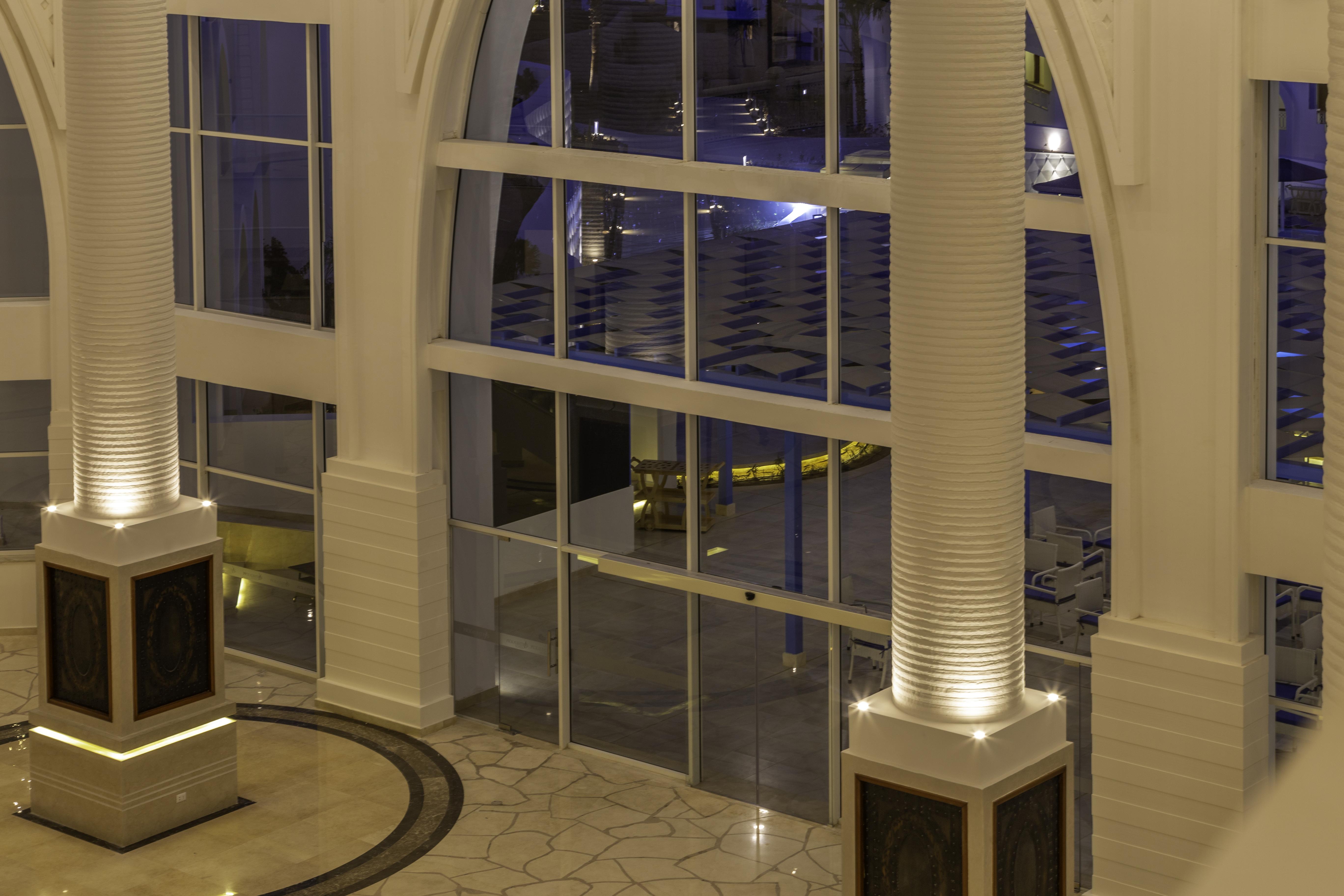 Pickalbatros Palace Sharm - "Aqua Park" Exterior foto The photo shows a spacious interior of a modern building, likely a hotel or a lobby. The scene features large glass windows that allow for a view of areas outside. There are elegant pillars within the space, illuminated from the base, creating a warm
