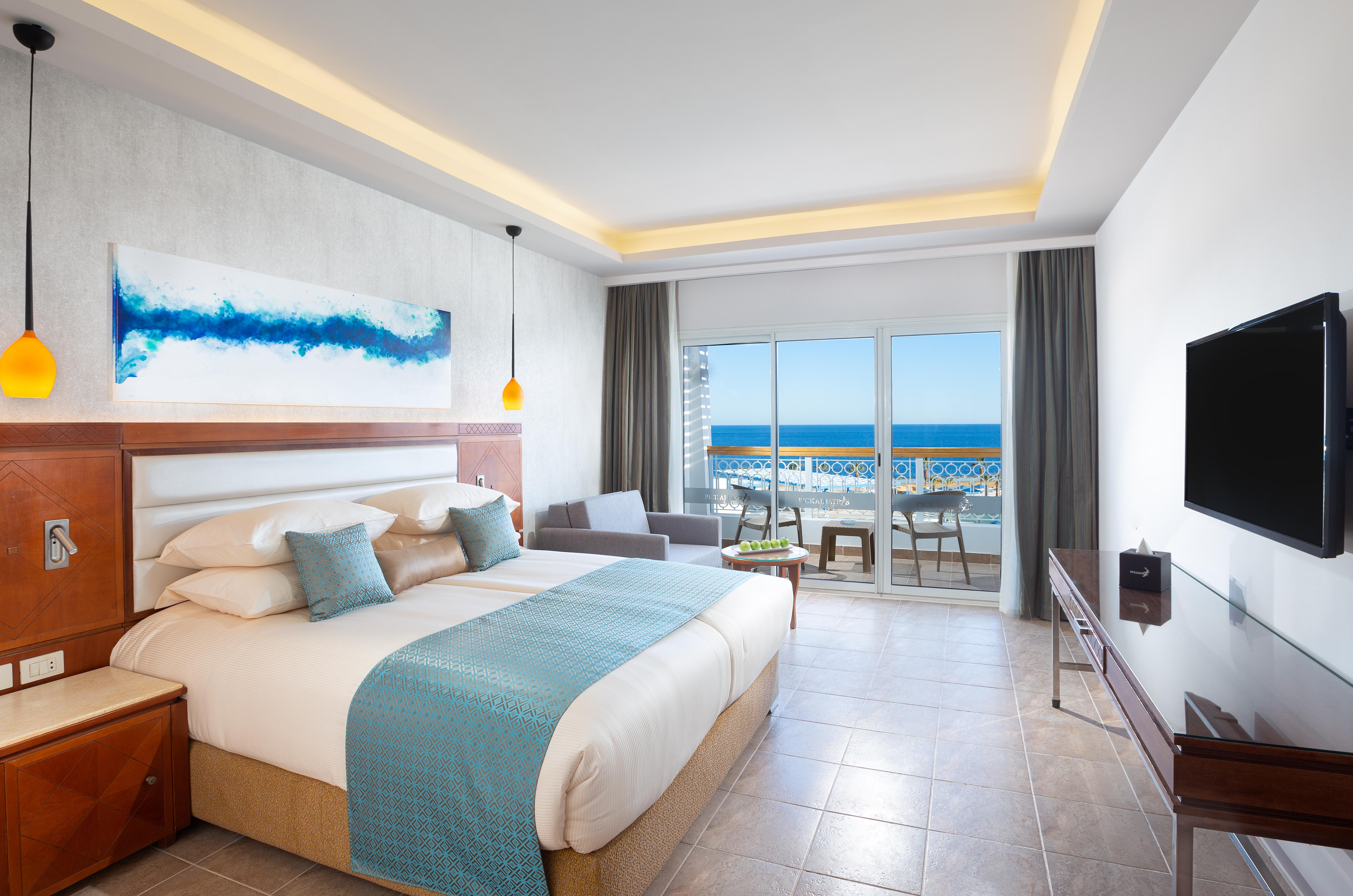 Pickalbatros Palace Sharm - "Aqua Park" Exterior foto The photo shows a modern hotel room with a large bed dressed in white linens and a light blue throw blanket. There is a piece of abstract artwork on the wall above the bed, featuring blue tones. The room has large windows with a view of the ocean, al