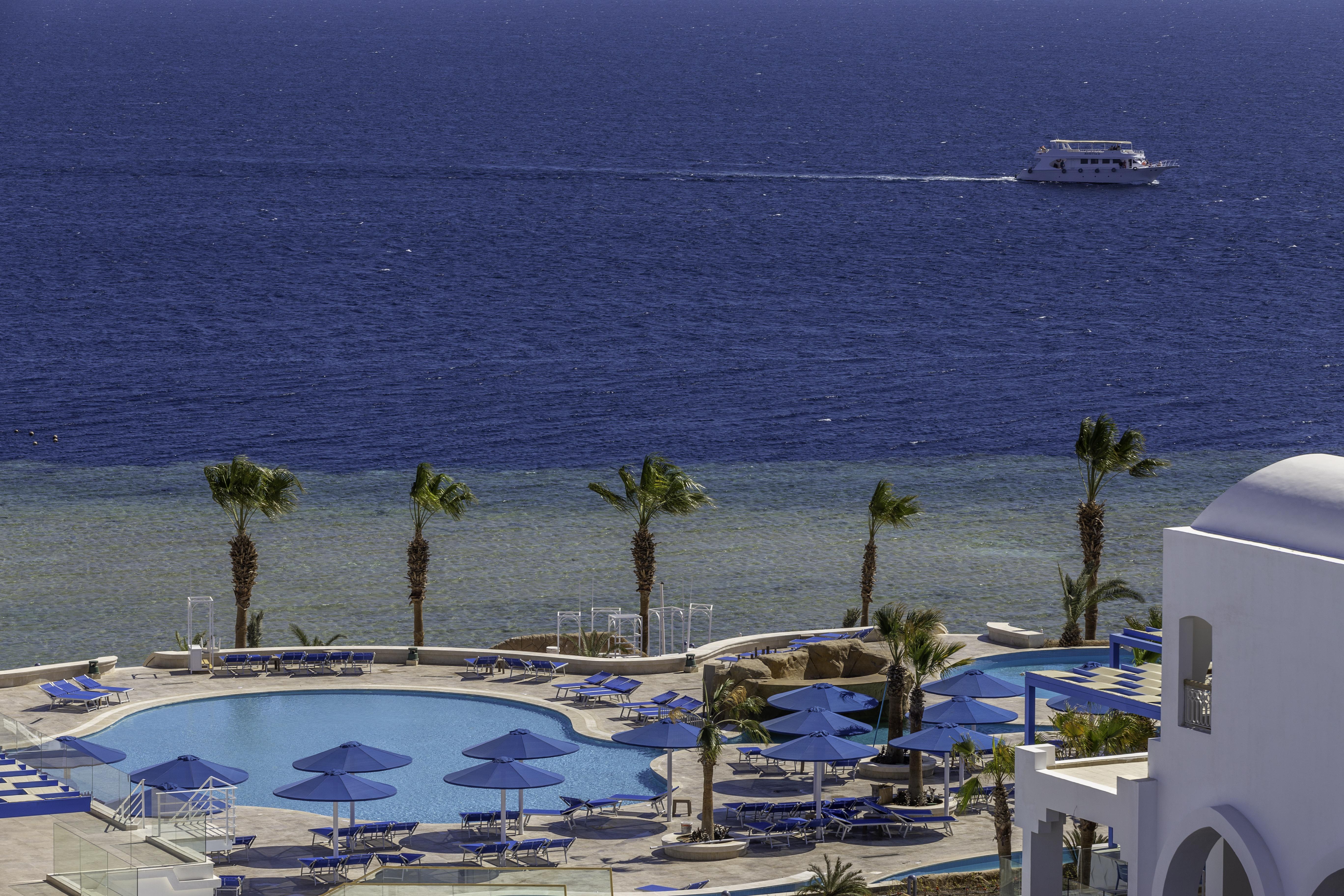 Pickalbatros Palace Sharm - "Aqua Park" Exterior foto The photo depicts a serene seaside view featuring a large swimming pool surrounded by lounge chairs and umbrellas. In the background, there is a calm sea with a boat cruising on the water. Palm trees line the edge of the pool area, adding to the trop