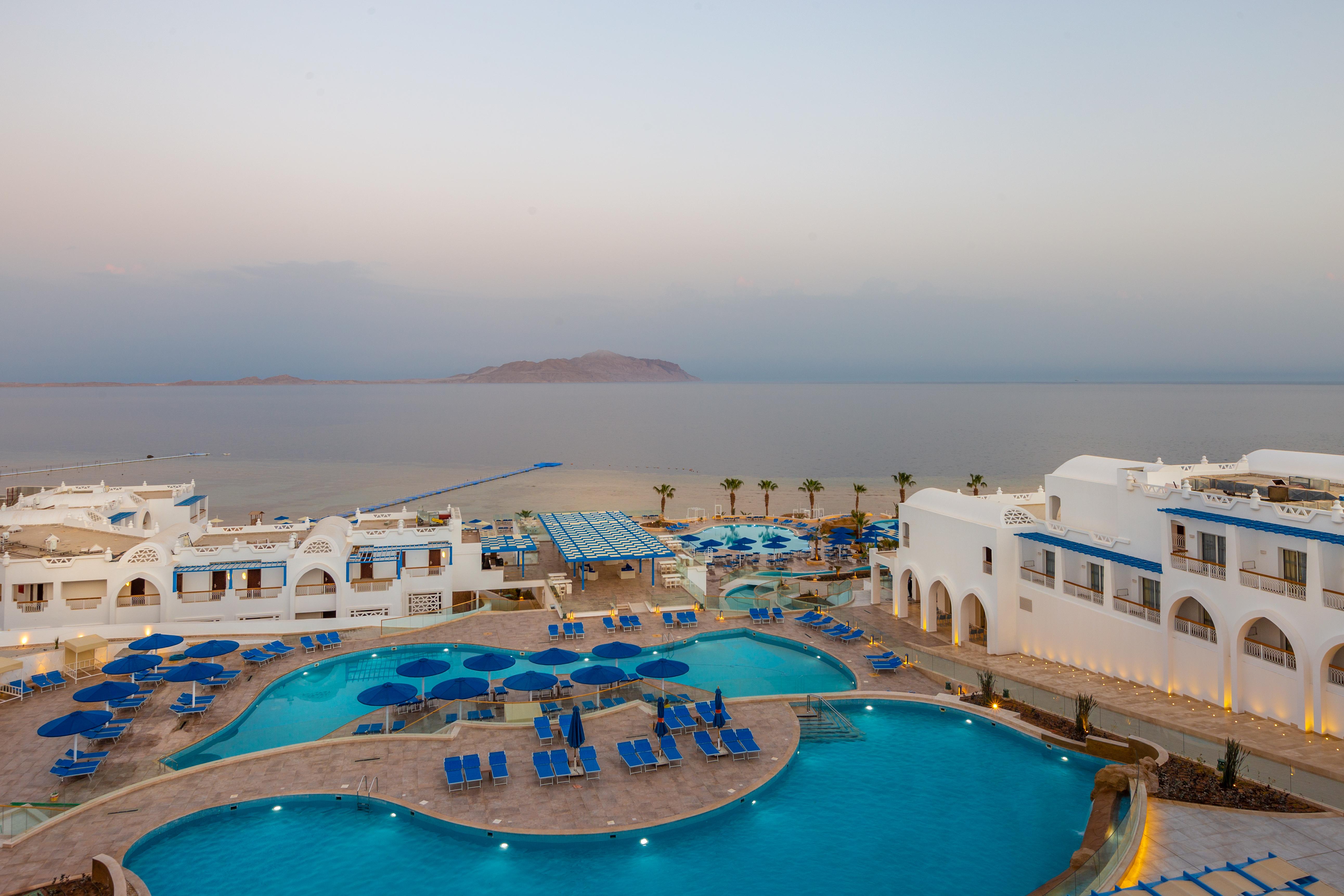 Pickalbatros Palace Sharm - "Aqua Park" Exterior foto The photo shows a resort area with expansive pools surrounded by lounge chairs. The pools are designed in a curvy shape, and there are numerous blue umbrellas scattered around. In the background, there is a calm sea stretching out towards a distant l