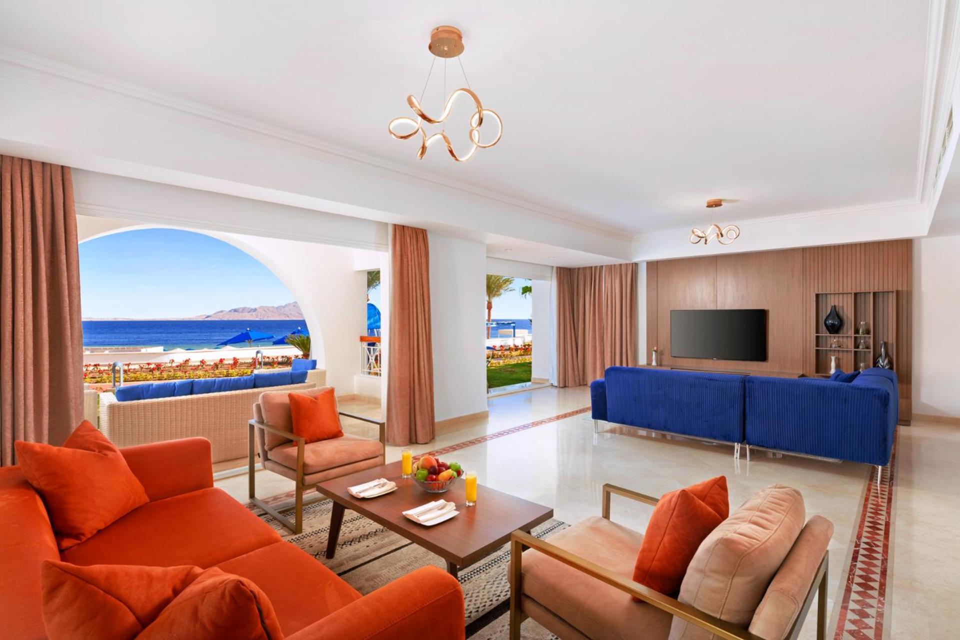 Pickalbatros Palace Sharm - "Aqua Park" Exterior foto The photo showcases a stylish and modern living space that features a bright and open layout. The room includes a cozy seating area with orange couches and a wooden coffee table, adorned with a fruit arrangement. Large windows provide a view of the o