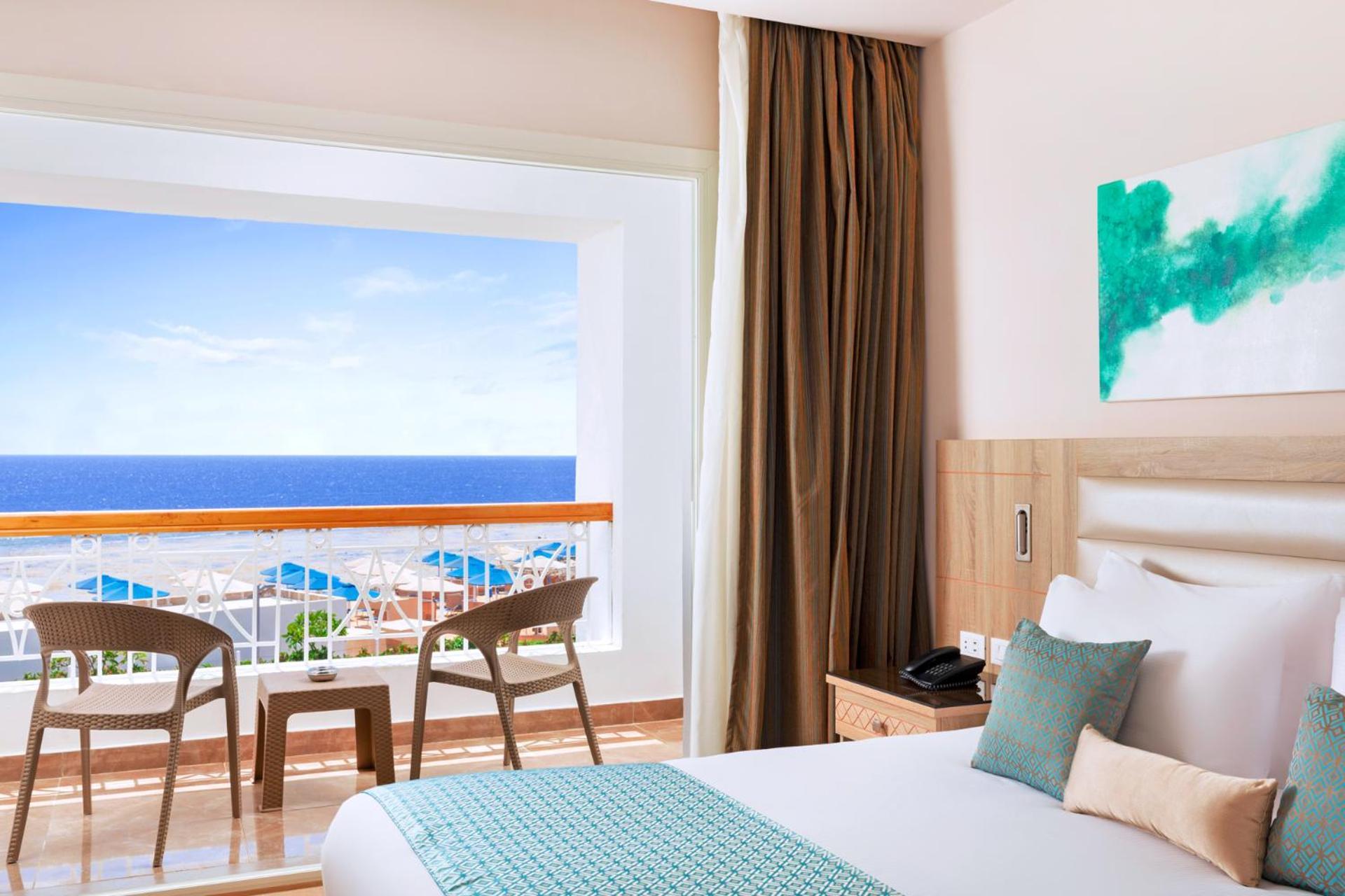 Pickalbatros Palace Sharm - "Aqua Park" Exterior foto The photo shows a modern hotel room featuring a large bed with white linens and a teal accent pillow. There is a window with a view of the ocean, framed by light curtains, which lets in natural light. In front of the window, there are two wooden chai