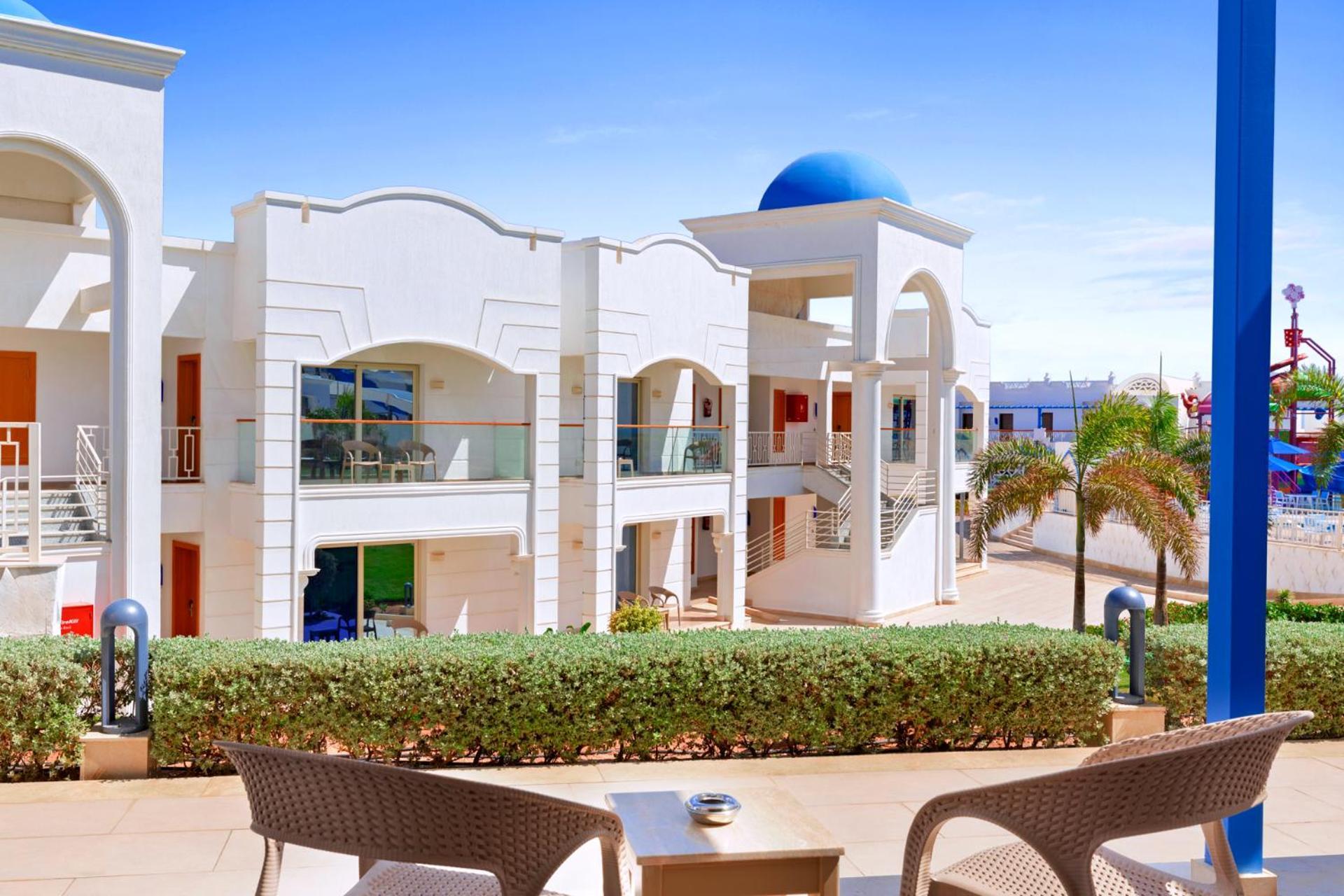 Pickalbatros Palace Sharm - "Aqua Park" Exterior foto The photo shows a modern architectural setting, likely at a resort or hotel. It features white buildings with decorative elements and balconies. The scene is bright with blue skies, and there are palm trees visible, adding a tropical vibe. In the for