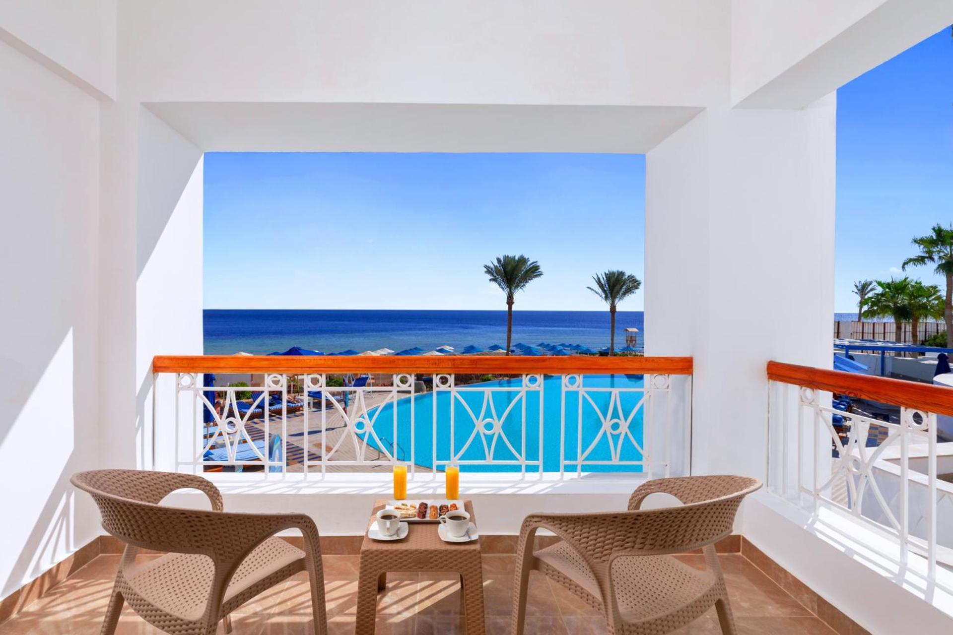 Pickalbatros Palace Sharm - "Aqua Park" Exterior foto The photo shows a bright and inviting balcony with two wicker chairs and a small table. On the table, there are two glasses of orange juice and a plate of food. The balcony overlooks a beautiful view of the ocean, with the deep blue water stretching 