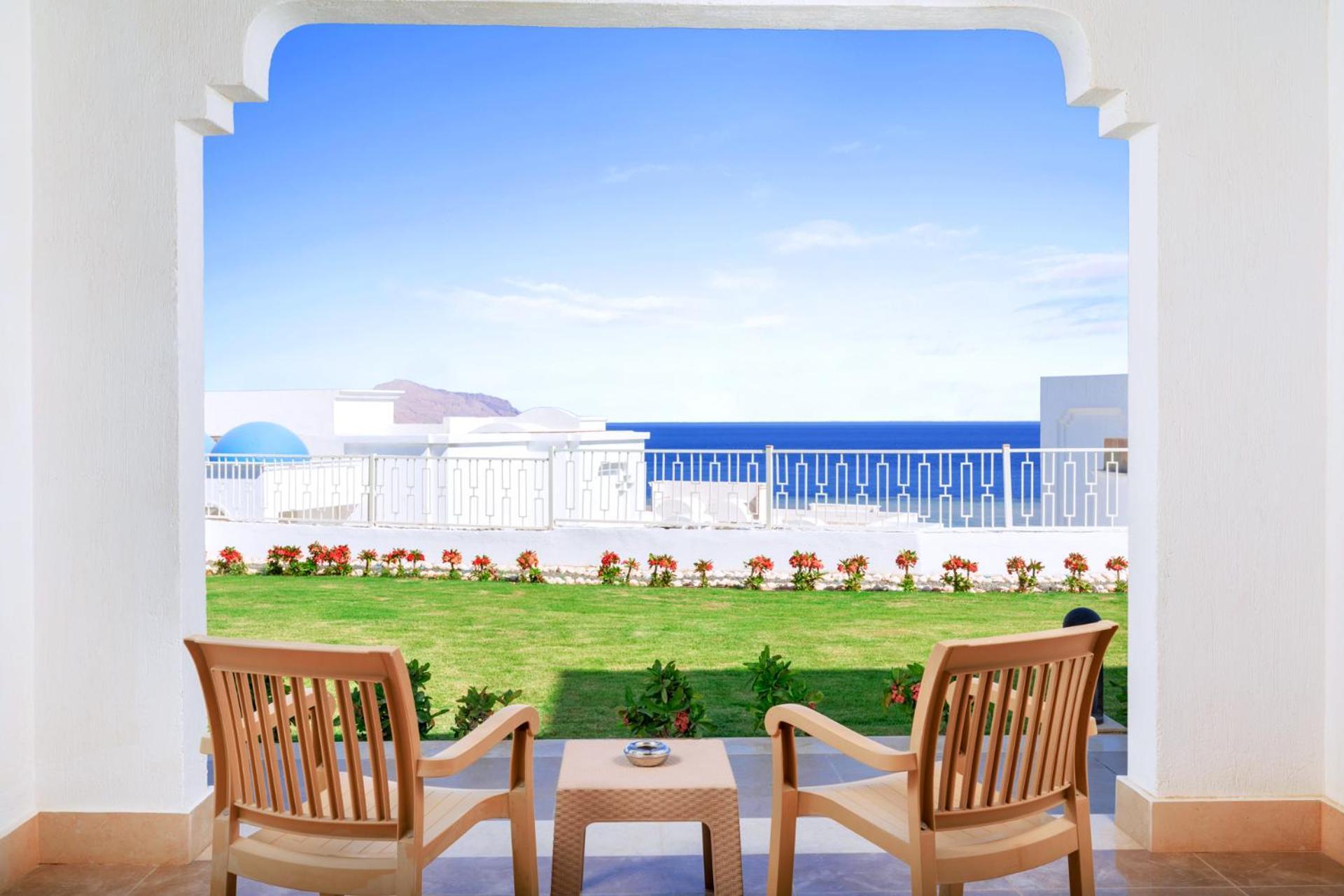 Pickalbatros Palace Sharm - "Aqua Park" Exterior foto The photo shows a scenic view from a terrace or balcony. In the foreground, there are two wooden chairs positioned around a small table. On the table, there is a decorative plant or vase. Beyond the seating area, a well-manicured lawn is visible, ado