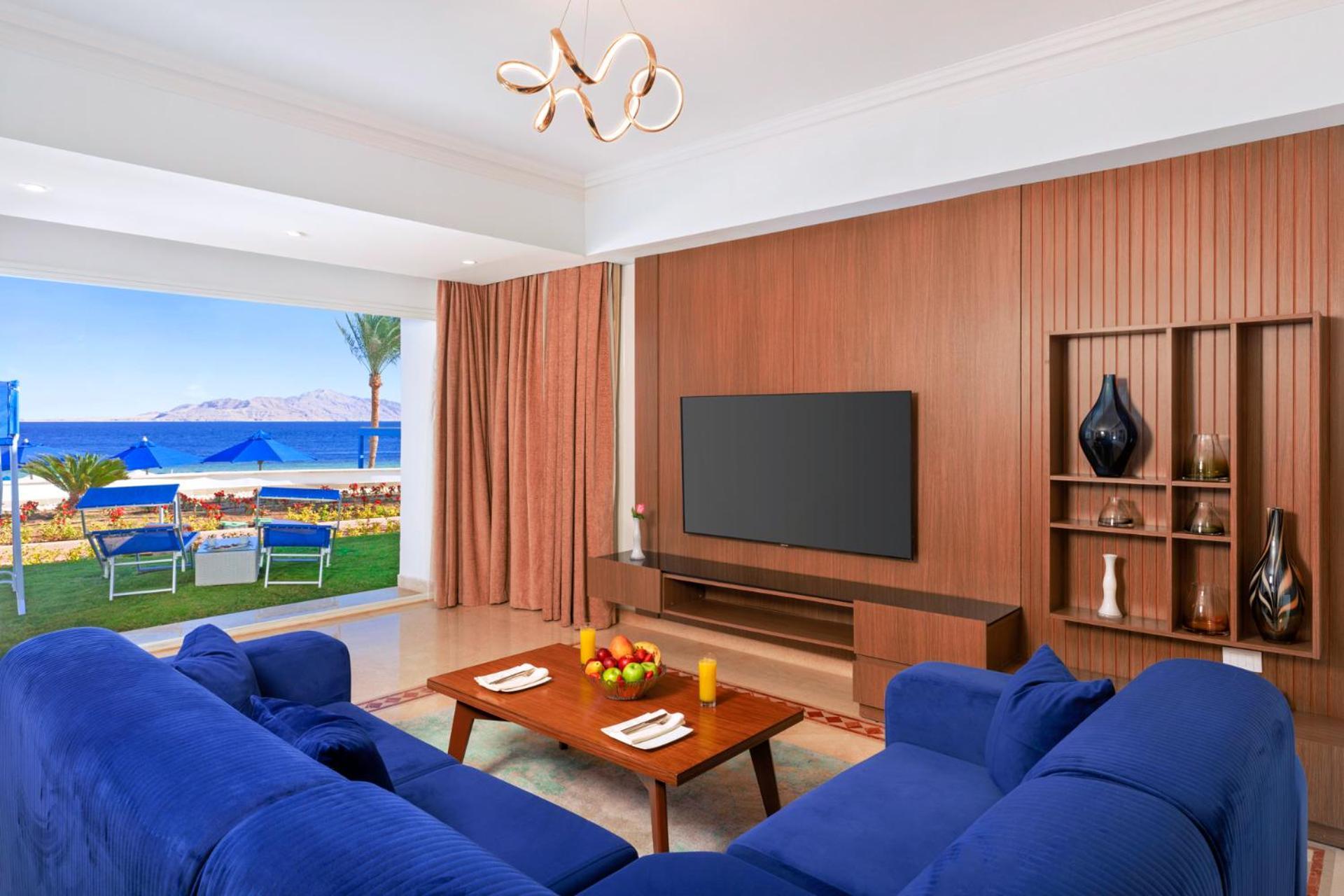 Pickalbatros Palace Sharm - "Aqua Park" Exterior foto The photo shows a modern living room with a stylish design. It features a blue sofa arranged around a wooden coffee table, which has some decorative items, including fruit and candles. A large flat-screen TV is mounted on a wooden wall panel. There a