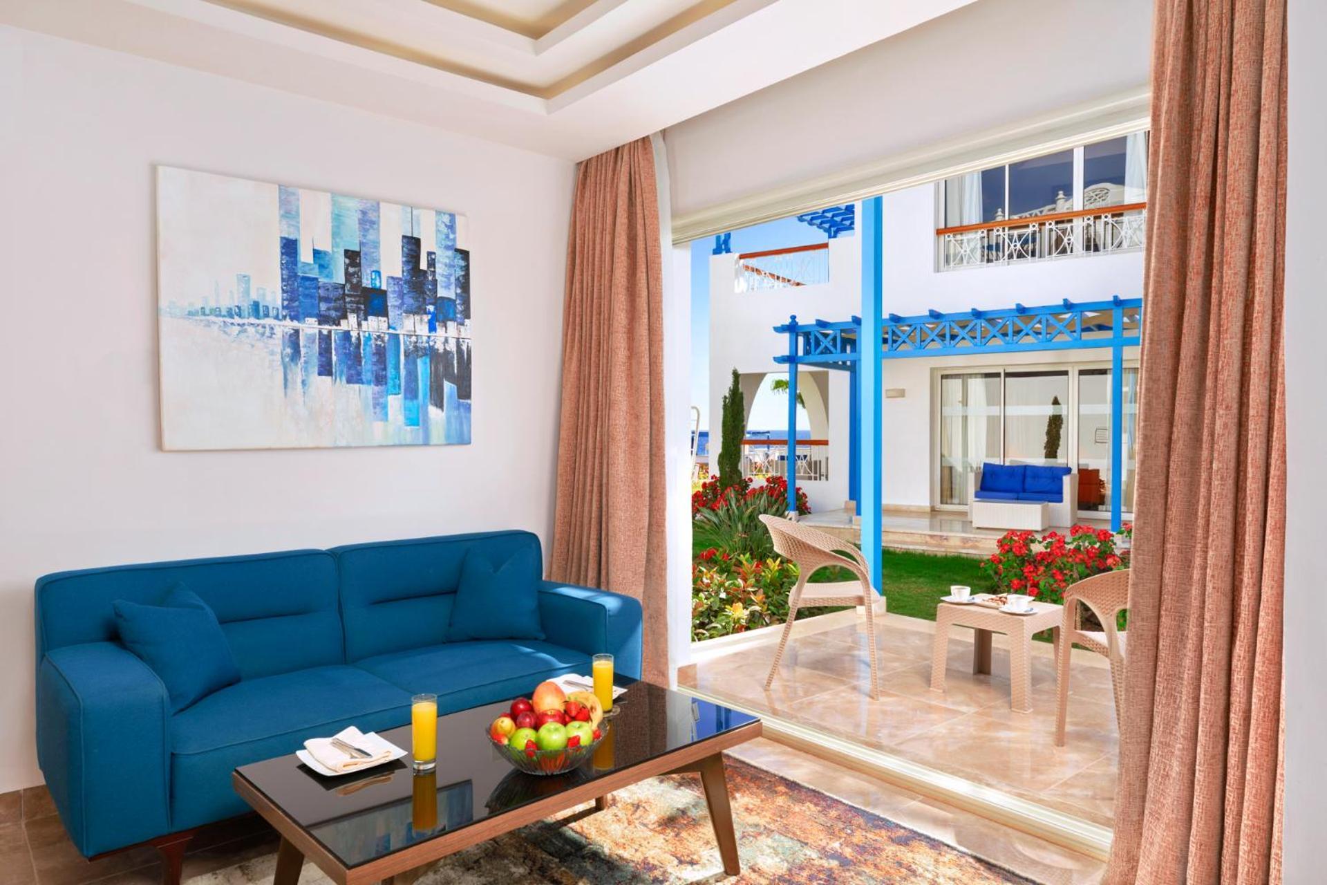 Pickalbatros Palace Sharm - "Aqua Park" Exterior foto The photo shows a bright and modern living space. There is a blue couch facing a glass door that leads to a garden area. In front of the couch, there's a dark coffee table with some fruit and candles on it. A large piece of abstract artwork in blues 