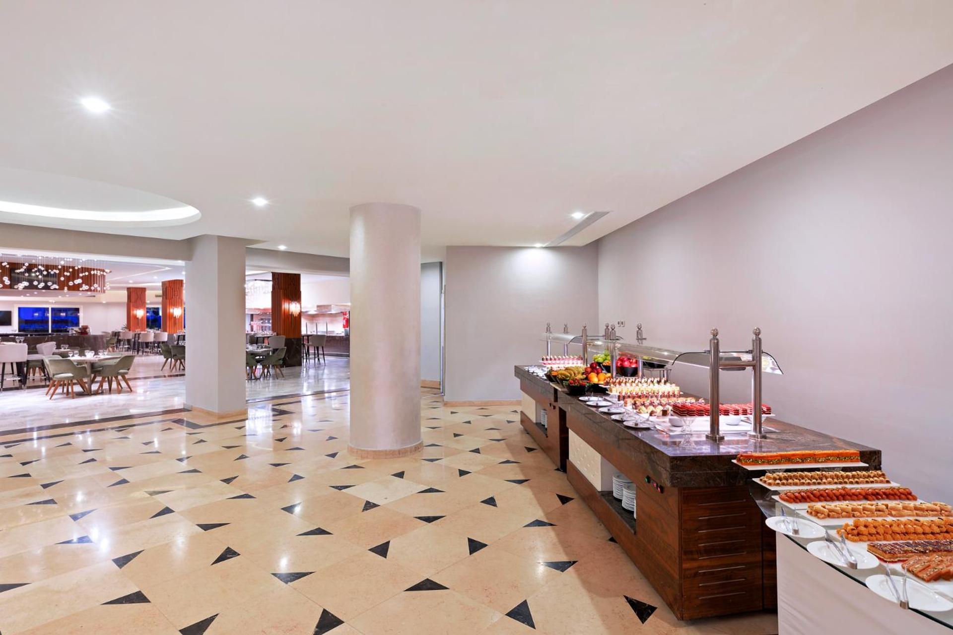 Pickalbatros Palace Sharm - "Aqua Park" Exterior foto The photo shows a modern dining area, likely a buffet or restaurant space. In the foreground, there's a wooden buffet table displaying various food items, including desserts. The floor has a decorative tile pattern, featuring a mix of light and darke